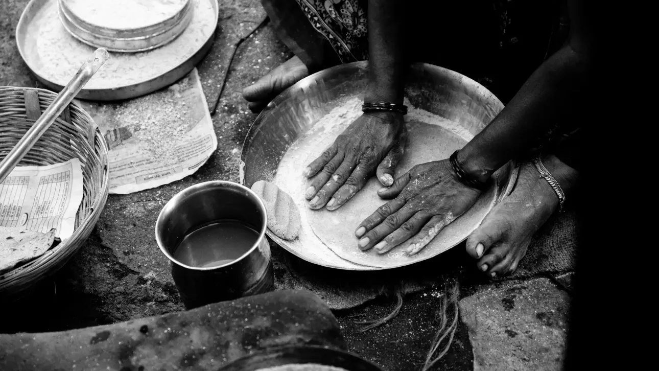 A woman sitting on the floor kneading flour in a metal pan to make roti